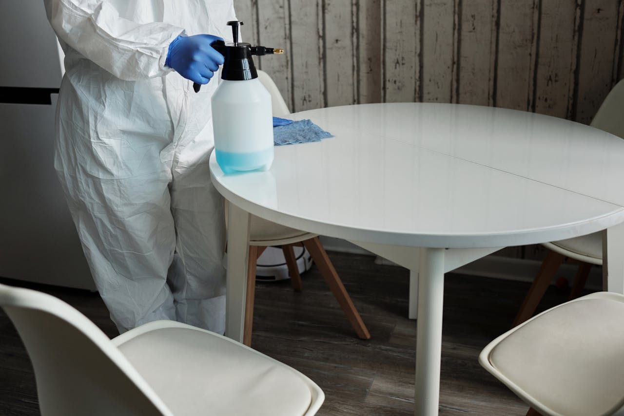 Person in protective suit disinfecting a white table indoors with a sprayer.